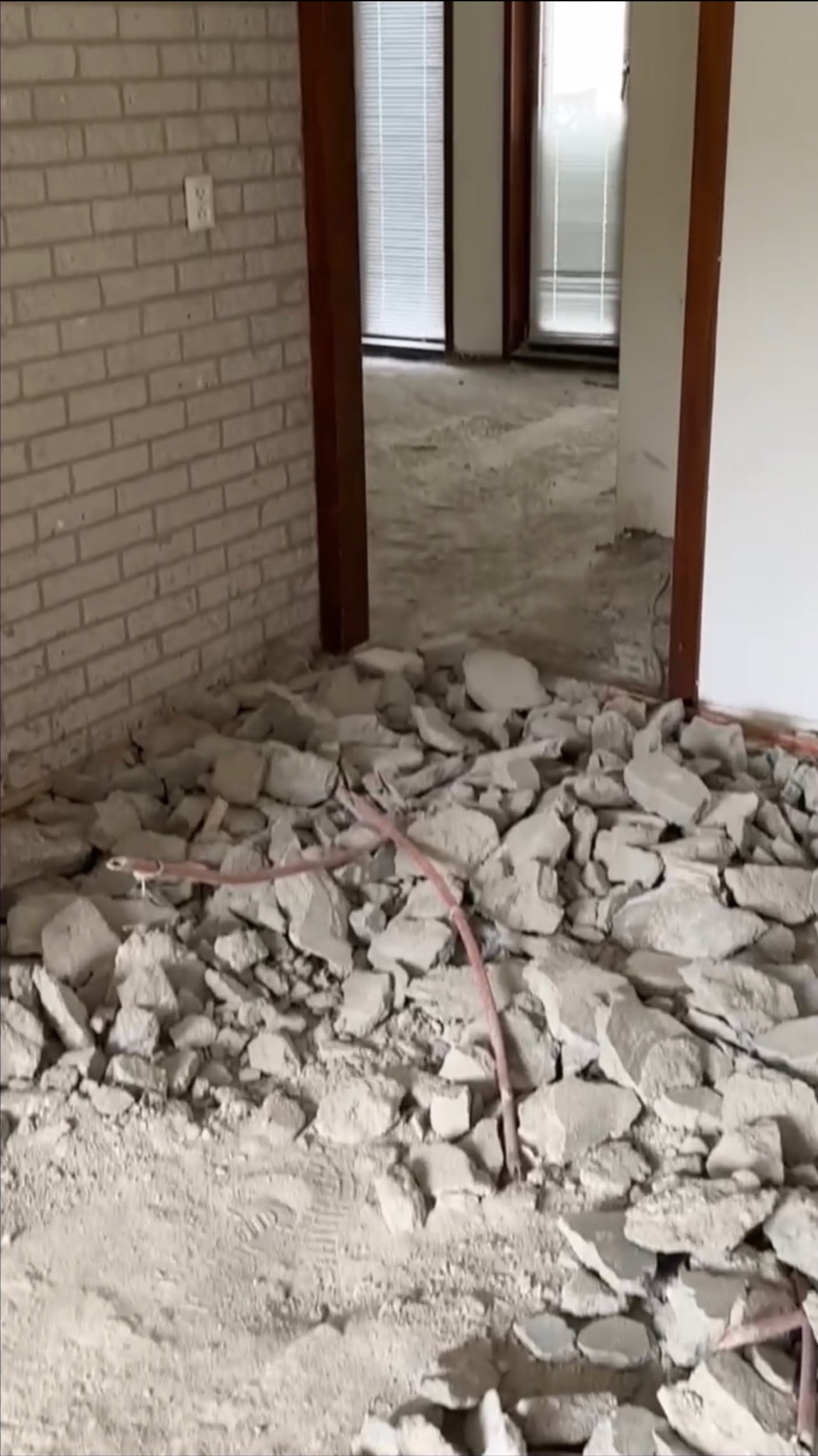 Wnętrze po demontażu posadzki, widoczne fragmenty gruzu, odsłonięta podłoga, fragment ściany z cegły oraz instalacja elektryczna w peszlu.