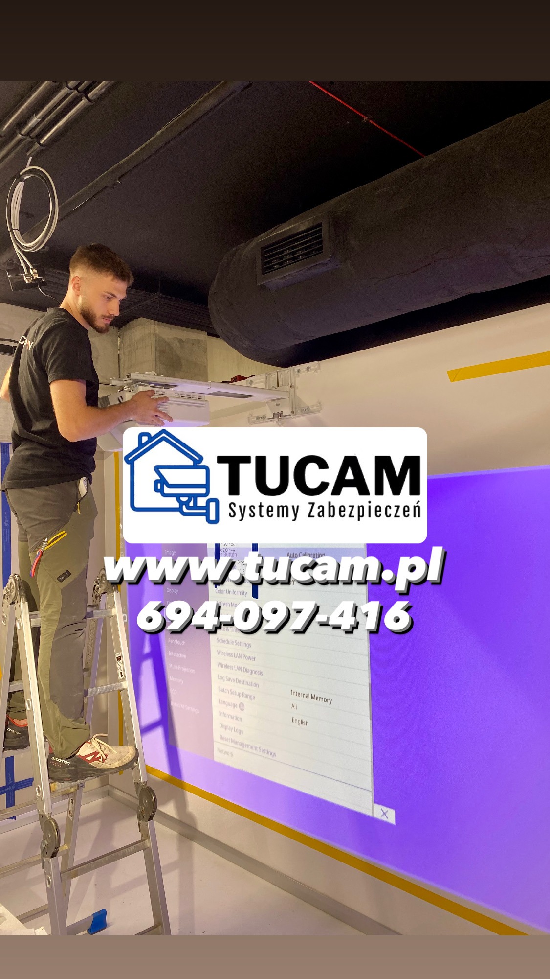 Instalator na drabinie montuje biały element systemu zabezpieczeń pod sufitem, widoczne logo firmy Tucam i ekran z ustawieniami systemu.