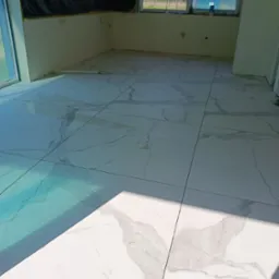 Wyłożona białymi płytkami podłoga w surowym wnętrzu z oknami zabezpieczonymi folią, widok na zewnątrz z zielenią i błękitem nieba.