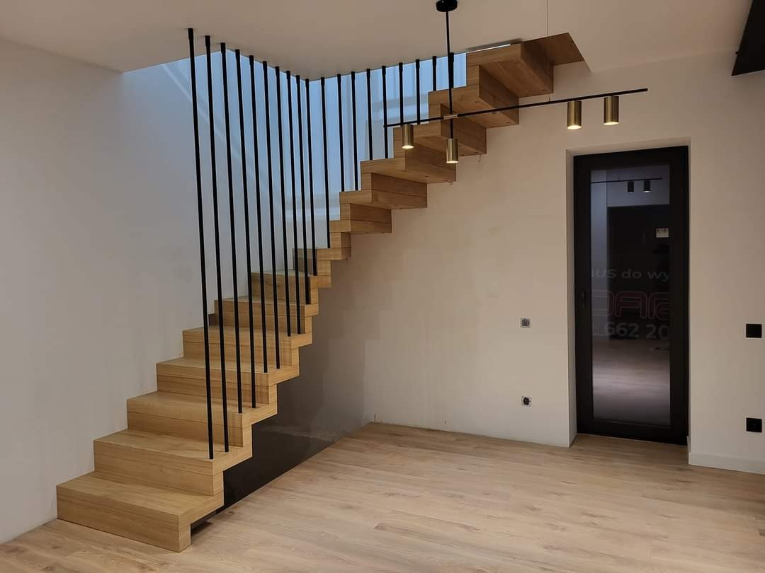 Nowoczesne, dębowe schody wspornikowe z czarnymi, pionowymi balustradami i oświetleniem punktowym, widoczne z perspektywy dolnej części biegu schodów.