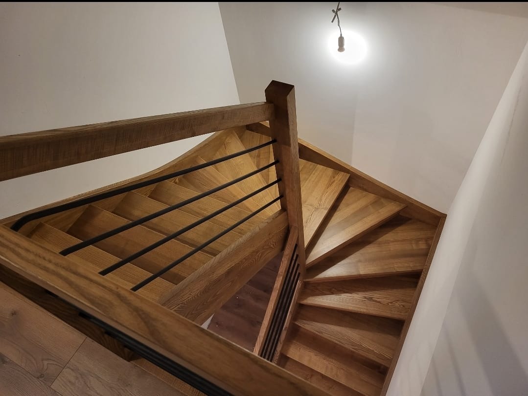 Widok z góry na drewniane schody spiralne z czarnymi metalowymi balustradami, oświetlone pojedynczą żarówką