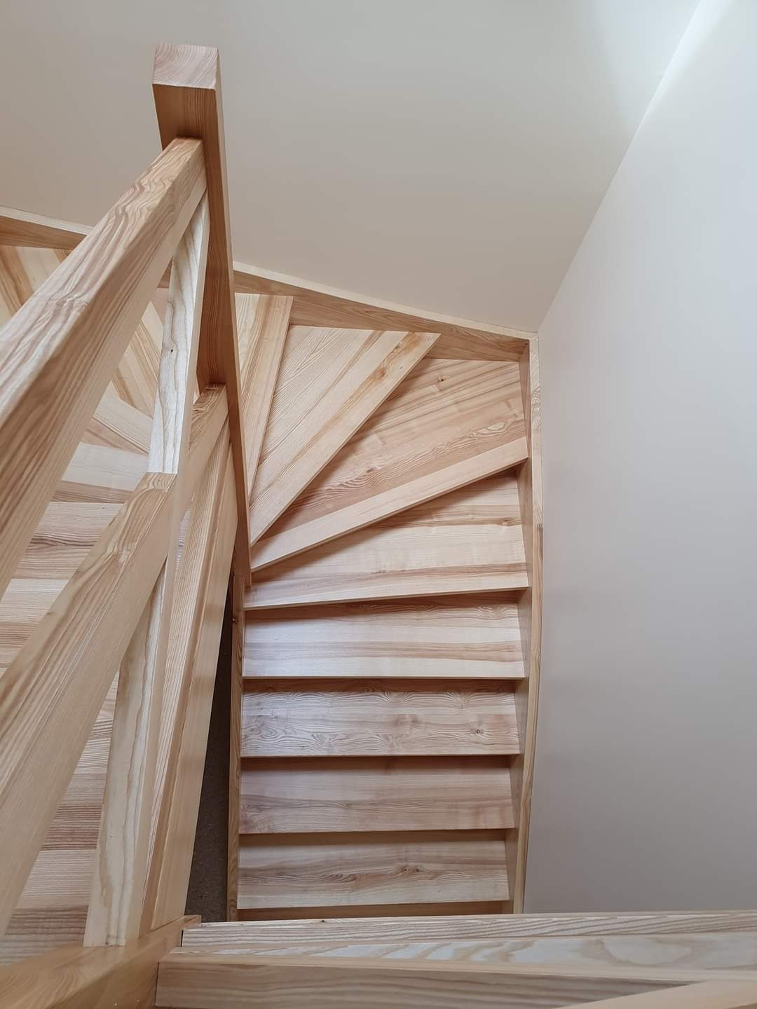 Drewniane schody spiralne z jasnego drewna, widok z góry, minimalistyczny design, białe ściany.