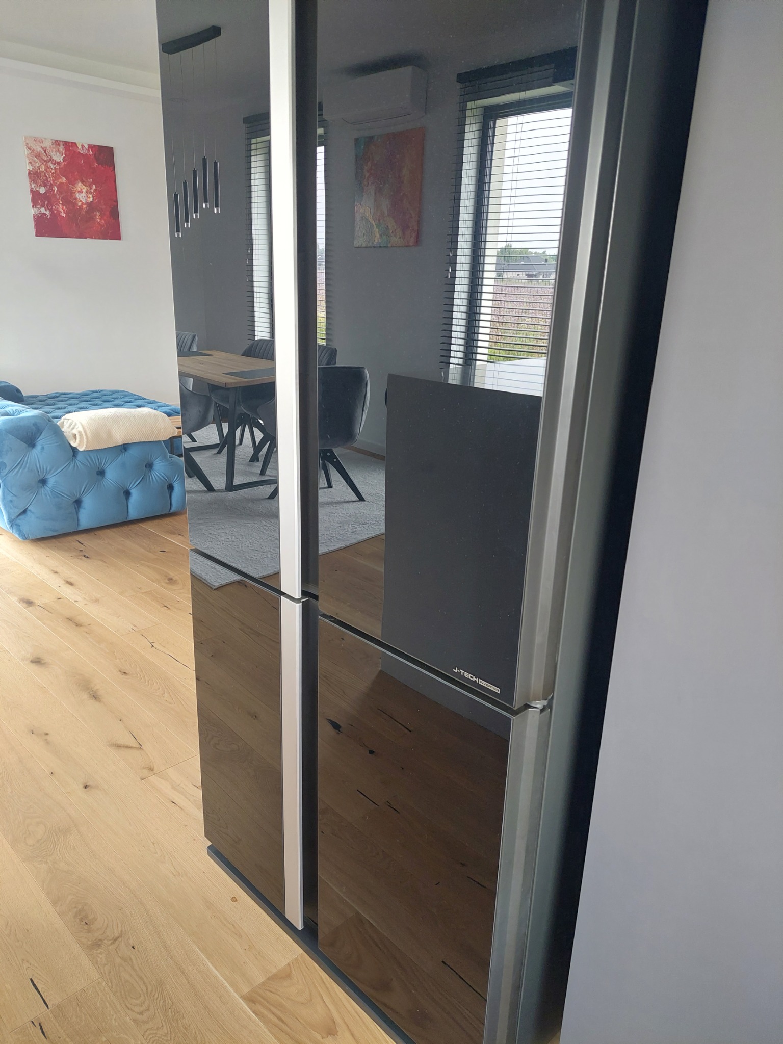 Nowoczesna kuchnia z drewnianą podłogą i lodówką z czarnym, lustrzanym frontem, w którym odbija się błękitna sofa i stół z krzesłami.