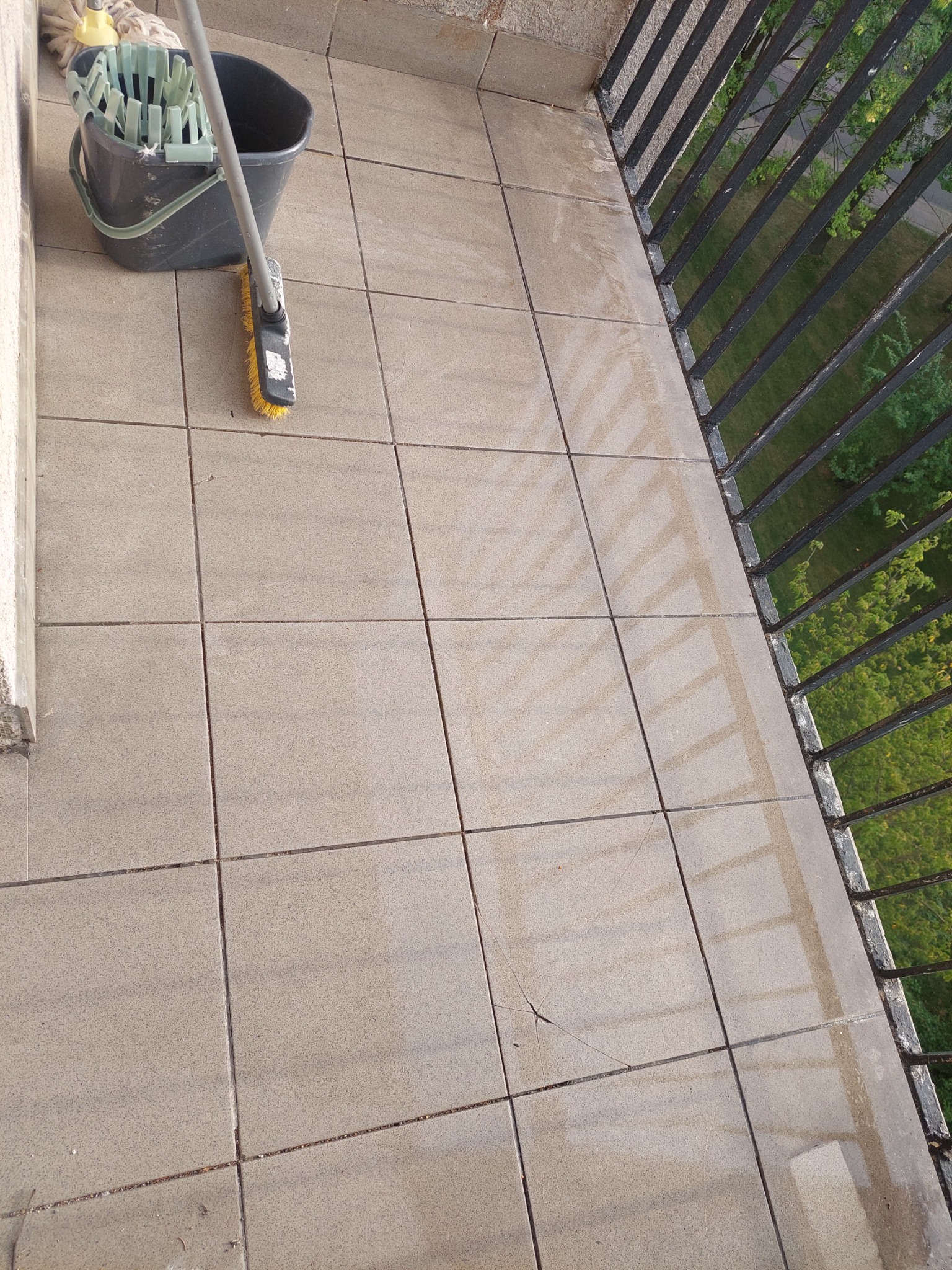 Mop i wiadro na mokrych, wyczyszczonych płytkach balkonu z widoczną szczeliną w jednej z płytek i czarną balustradą z widokiem na zieleń.