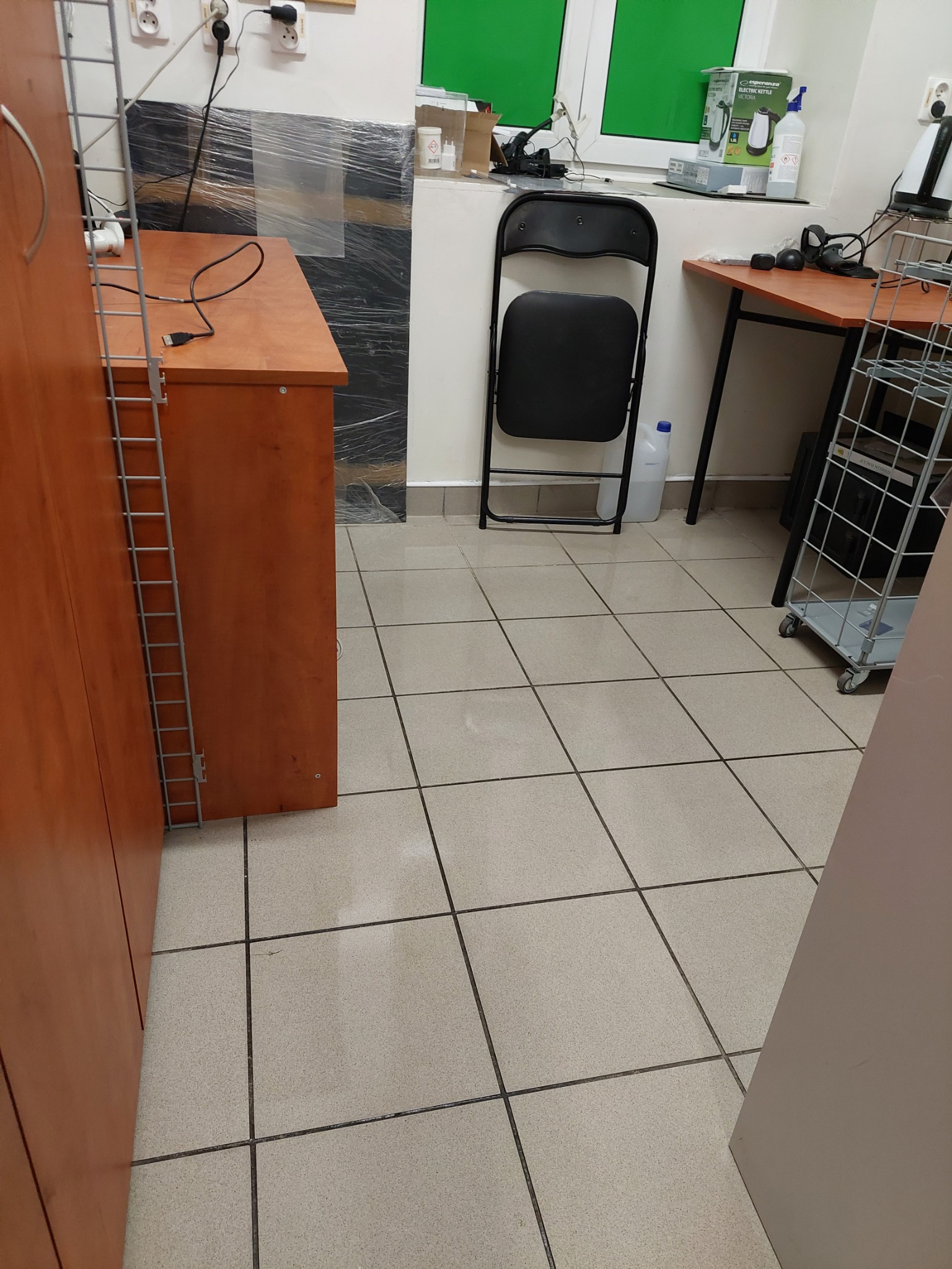 Wnętrze biura z widoczną podłogą z płytek, biurkiem z elektroniką, krzesłem składanym i regałem na kółkach, w tle okno z zieloną roletą.