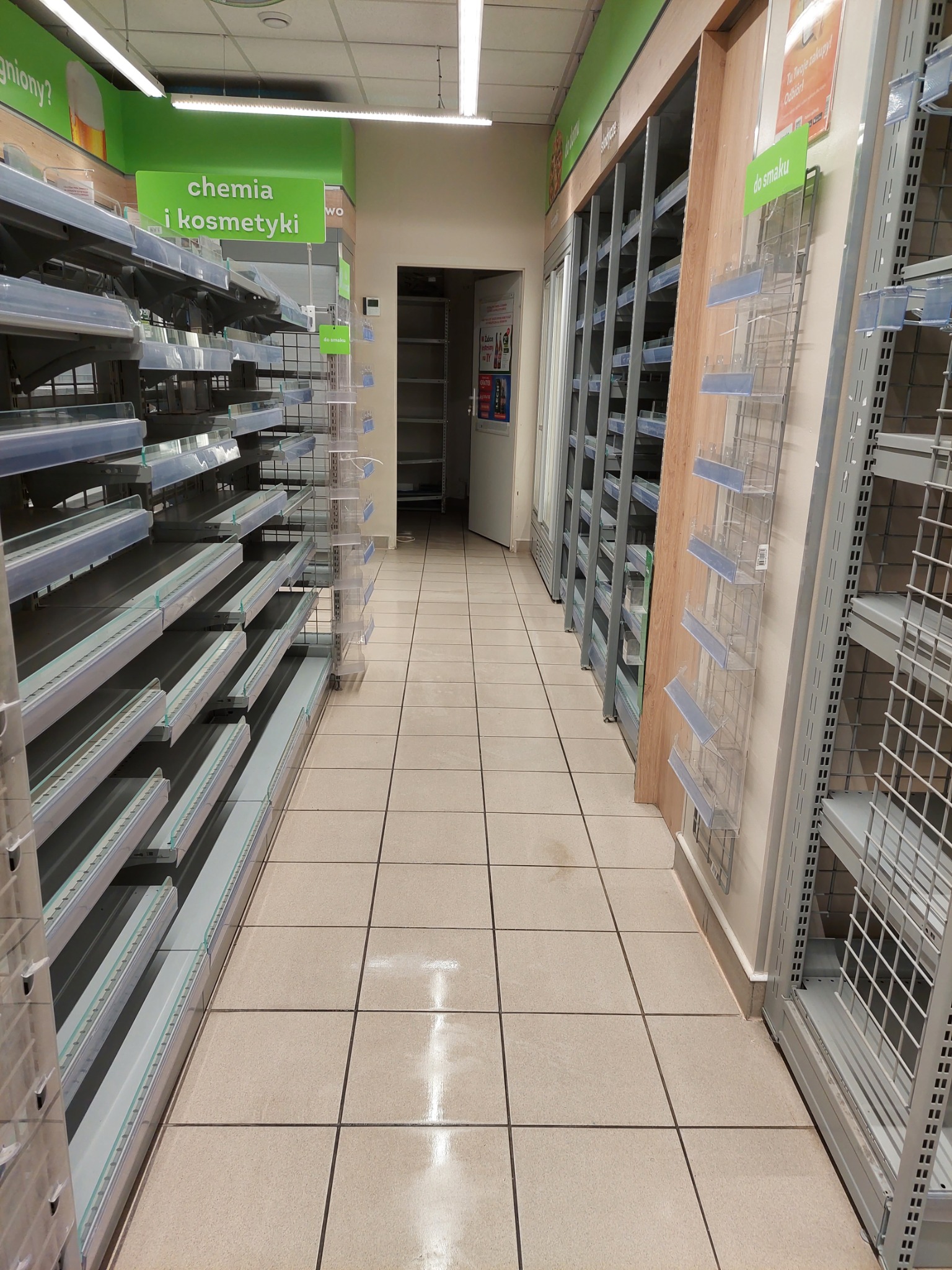 Pusta alejka w sklepie z półkami na chemię i kosmetyki, beżowe płytki podłogowe odbijają światło.