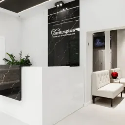 Nowoczesne wnętrze kliniki stomatologicznej: biała recepcja z czarnym marmurowym akcentem, logo 'dentusplus' na czarnym panelu, białe fotele dla pacjentów i telewizory w tle.