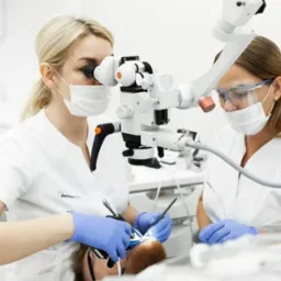 Lekarz stomatolog w trakcie zabiegu z użyciem mikroskopu stomatologicznego, asystuje mu drugi lekarz, obaj w maseczkach i rękawiczkach, widoczny pacjent na fotelu dentystycznym.