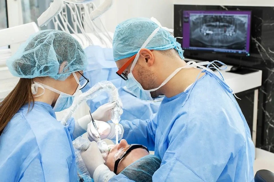 Stomatolog w trakcie zabiegu, asystentka przy głowie pacjenta, widoczny monitor z prześwietleniem zębów, oświetlenie stomatologiczne.