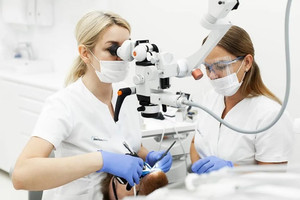 Lekarz stomatolog w trakcie zabiegu z użyciem mikroskopu stomatologicznego, asystuje mu drugi lekarz, obaj w maseczkach i rękawiczkach, widoczny pacjent na fotelu dentystycznym.