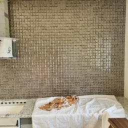 Ściana w kuchni wyłożona błyszczącymi, szarymi płytkami 3D z efektem mozaiki, zabezpieczona białą płachtą i wzorzystą szmatką, widoczne fragmenty szafek kuchennych i oświetlenia sufitowego.