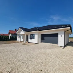 Nowoczesny, parterowy dom z białą elewacją, grafitowym dachem i bramą garażową, otoczony jasnym żwirem, w słoneczny dzień.
