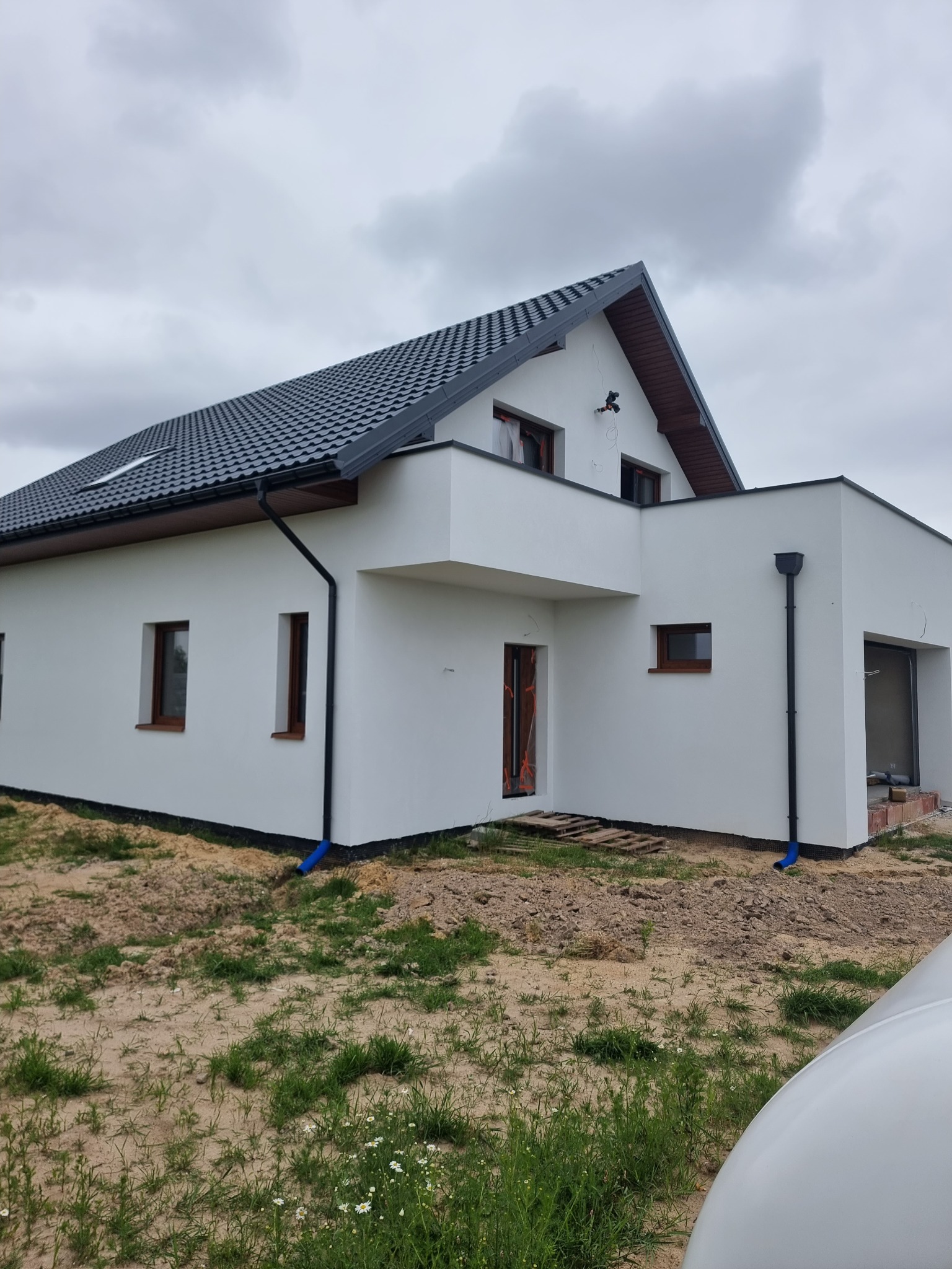 Nowoczesny dom jednorodzinny z białą elewacją, ciemną dachówką i brązowymi oknami, widok z zewnątrz w pochmurny dzień, częściowo wykończony teren wokół budynku.
