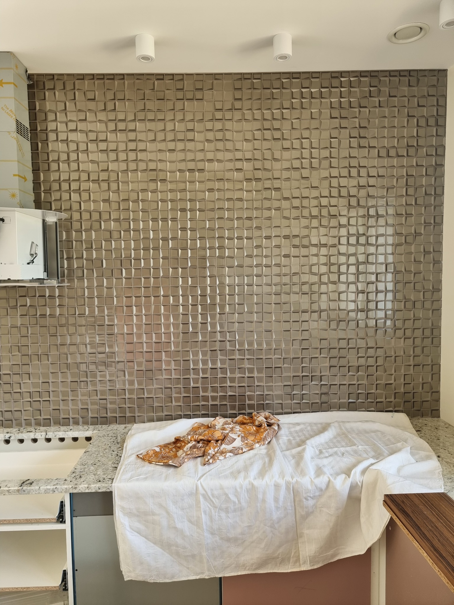 Ściana w kuchni wyłożona błyszczącymi, szarymi płytkami 3D z efektem mozaiki, zabezpieczona białą płachtą i wzorzystą szmatką, widoczne fragmenty szafek kuchennych i oświetlenia sufitowego.