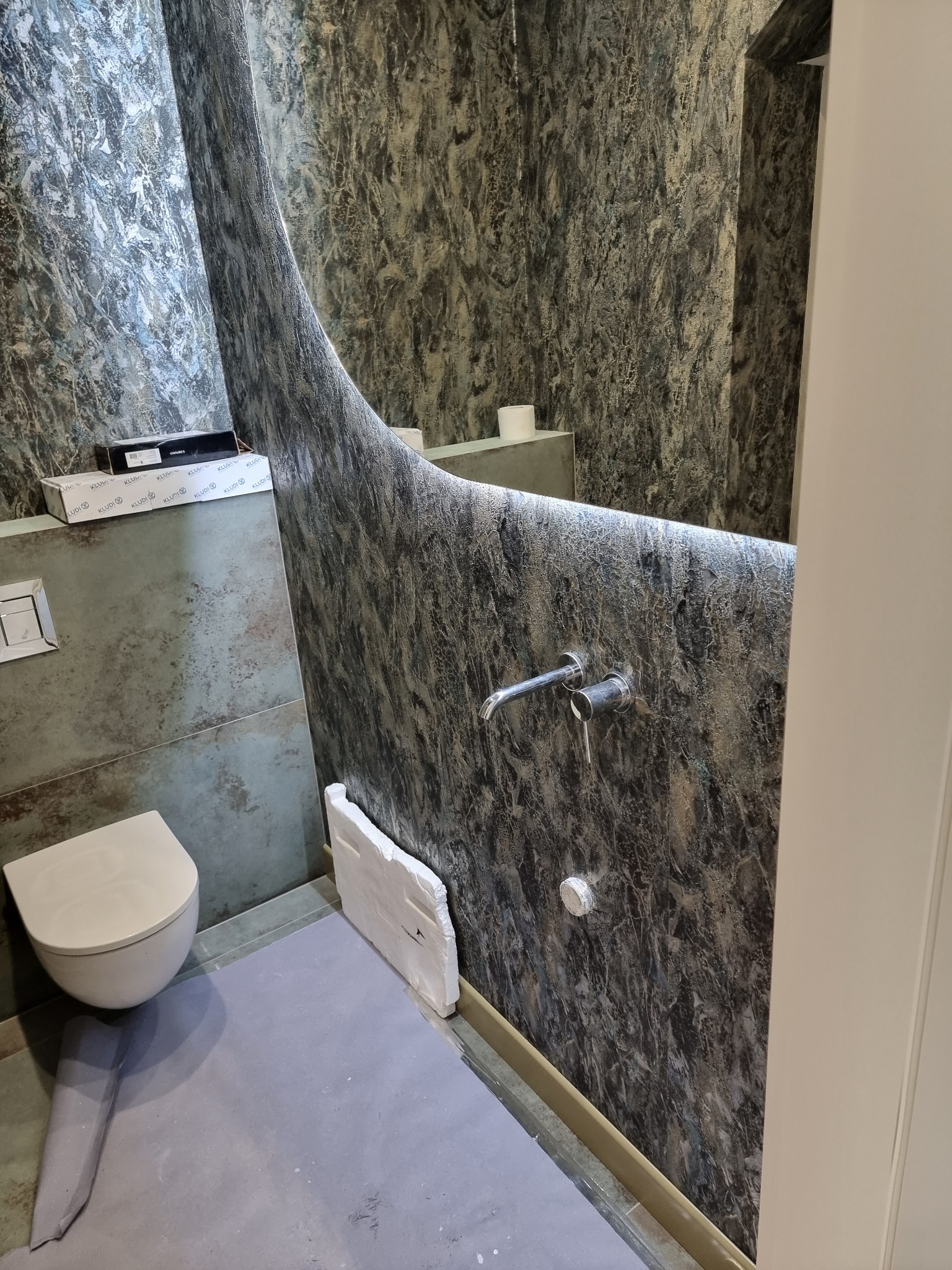 Niedokończona łazienka z toaletą podwieszaną, surowymi ścianami imitującymi marmur i zabezpieczoną podłogą, widoczne opakowania po armaturze.