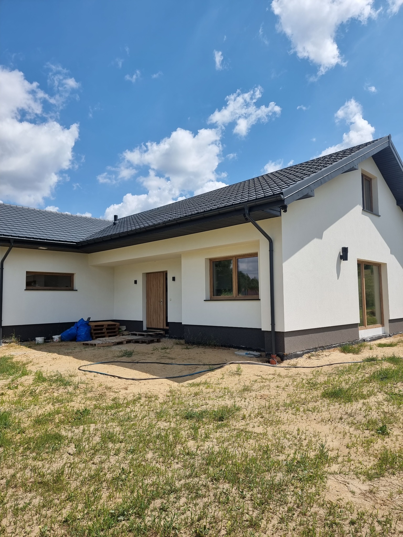 Nowoczesny dom jednorodzinny z białą elewacją i grafitowym dachem, widok z zewnątrz, na trawniku widoczne ślady piasku i pozostałości po budowie.