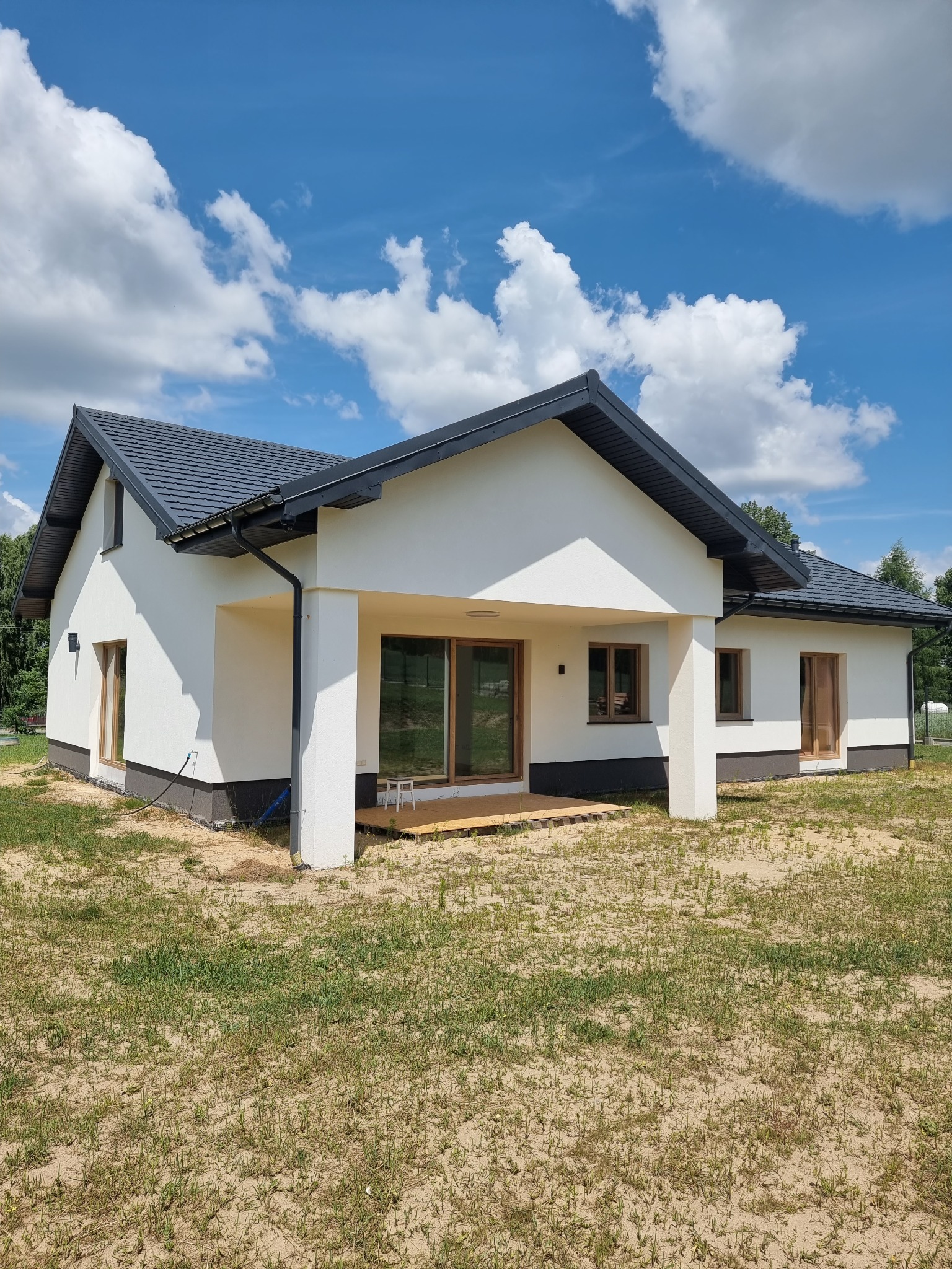 Nowoczesny dom jednorodzinny z białą elewacją, grafitowym dachem i drewnianymi oknami, widoczny z boku w słoneczny dzień z błękitnym niebem i białymi chmurami w tle.
