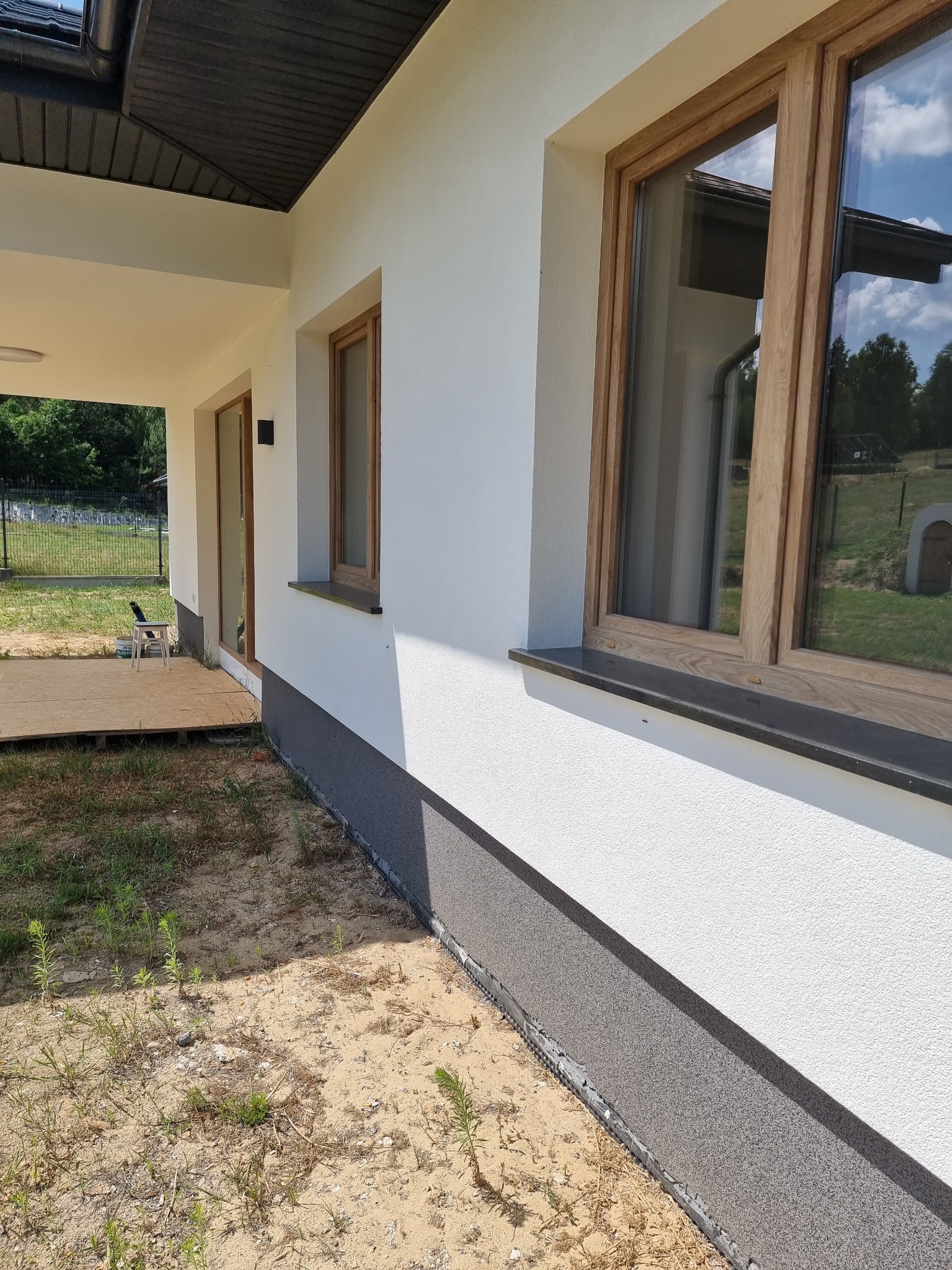 Fragment elewacji domu jednorodzinnego z drewnianą stolarką okienną i szarą podmurówką, widok z zewnątrz na niezagospodarowany teren.