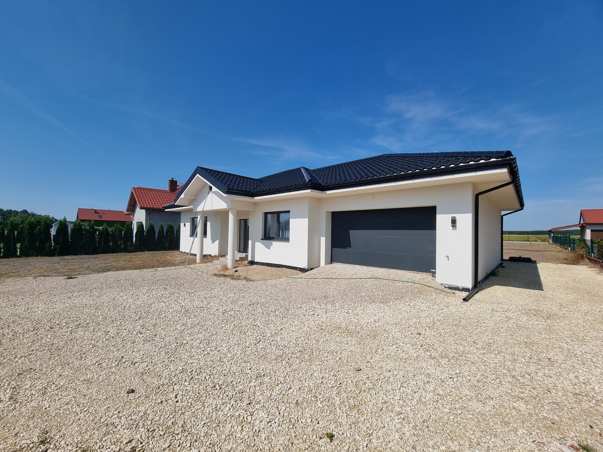 Nowoczesny, parterowy dom z białą elewacją, grafitowym dachem i bramą garażową, otoczony jasnym żwirem, w słoneczny dzień.