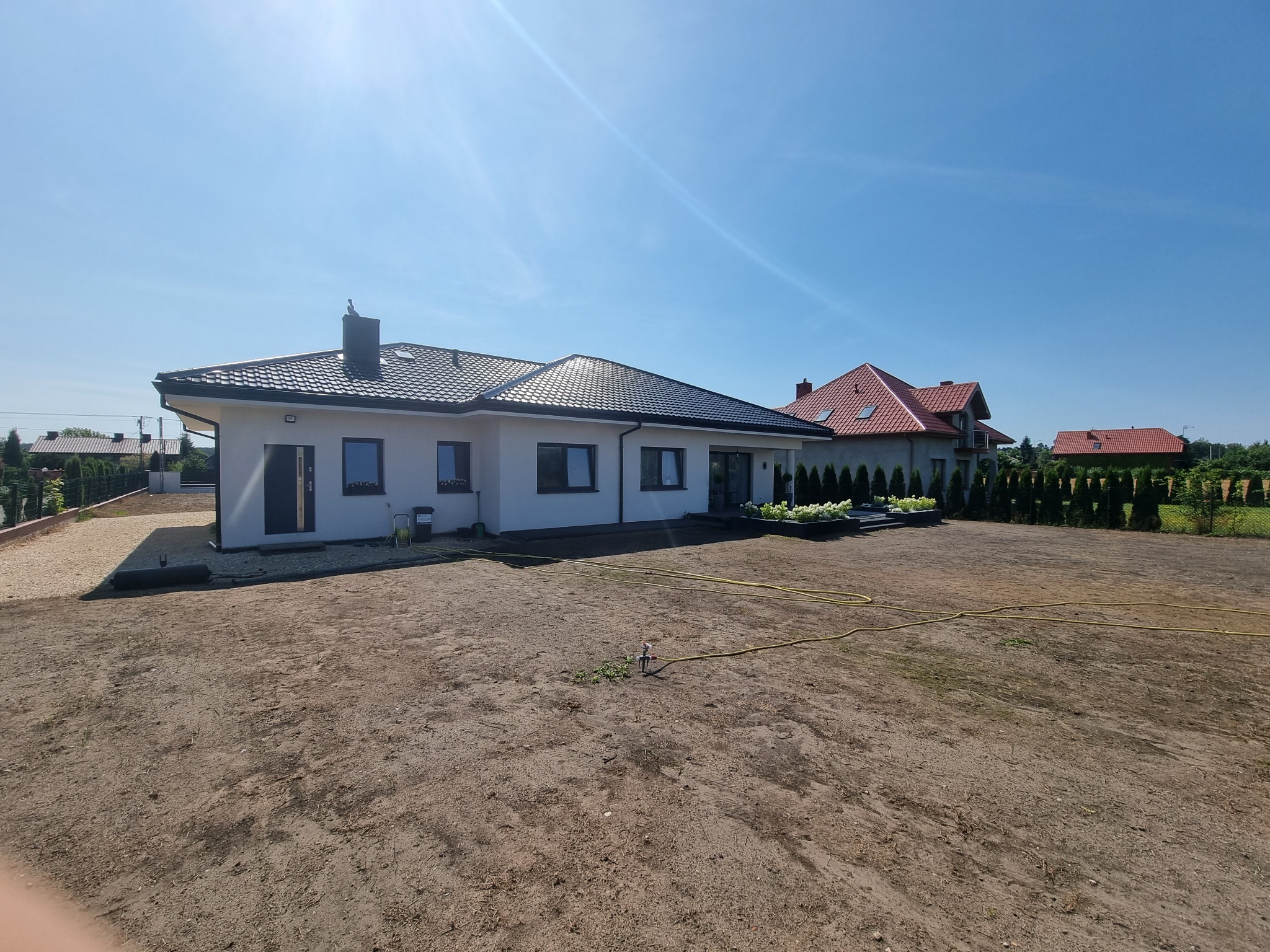 Nowoczesny, parterowy dom z białą elewacją i ciemną dachówką, widok z ogrodu z systemem nawadniania, błękitne niebo.