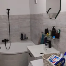 Jasna łazienka z wanną obudowaną mozaiką, umywalką z czarną armaturą, ośmiokątnym lustrem i pralką z koszykiem na środki czystości.