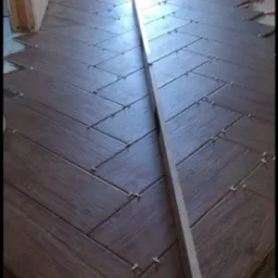 Układanie ciemnobrązowych płytek podłogowych w jodełkę, z użyciem poziomicy i krzyżyków dystansowych, widoczna warstwa kleju na podłożu.