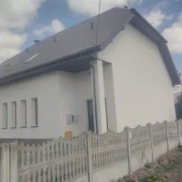 Świeżo pomalowany dom jednorodzinny z szarą dachówką, widok zza betonowego ogrodzenia, biała elewacja, rynna w kolorze dachu.