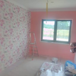 ECO-HOUSE Mateusz Kaliński - Pokój w trakcie remontu: ściana z tapetą w kwiaty, różowe ściany, drabina, okno z widokiem na zieleń, materiały i narzędzia malarskie na podłodze.