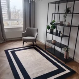 Jasny dywan z ciemnym obramowaniem na parkiecie w pokoju z czarnym regałem i fotelem w pepitkę.