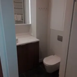 Nowoczesna łazienka z wiszącą toaletą, szafką pod umywalką z ciemnego drewna i podświetlanym lustrem z wbudowaną szafką, widok na grzejnik drabinkowy i czarne płytki podłogowe.