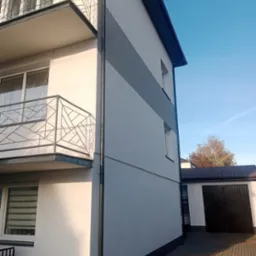 Dwupiętrowy budynek po remoncie elewacji, z balkonami z metalowymi balustradami o geometrycznym wzorze, pomalowany na biało z szarym pasem, widok z dołu na bocznej elewacji, rynna w kolorze...