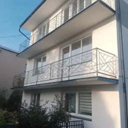 Dwupiętrowy dom z białymi ścianami i balkonami z geometrycznymi balustradami, oświetlony porannym słońcem, z zielenią u podstawy.