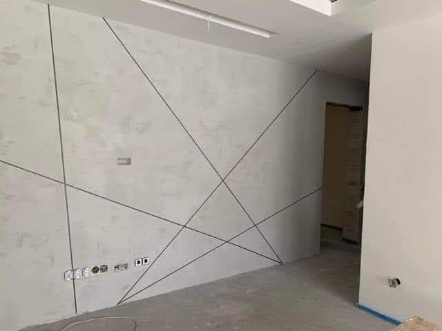 Surowa ściana wewnętrzna z zaznaczonymi liniami podziału, widoczne gniazdka elektryczne i fragment podłogi w trakcie remontu.