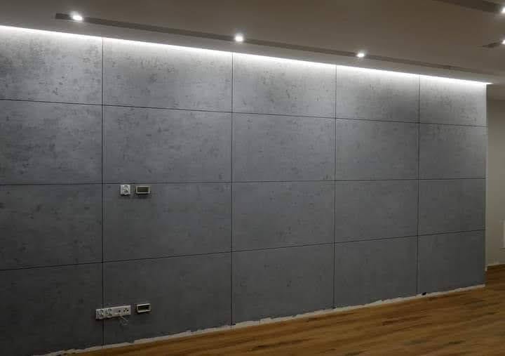 Ściana wykończona szarymi płytami imitującymi beton, z widocznymi gniazdkami elektrycznymi i włącznikami, podświetlona listwą LED podsufitową.