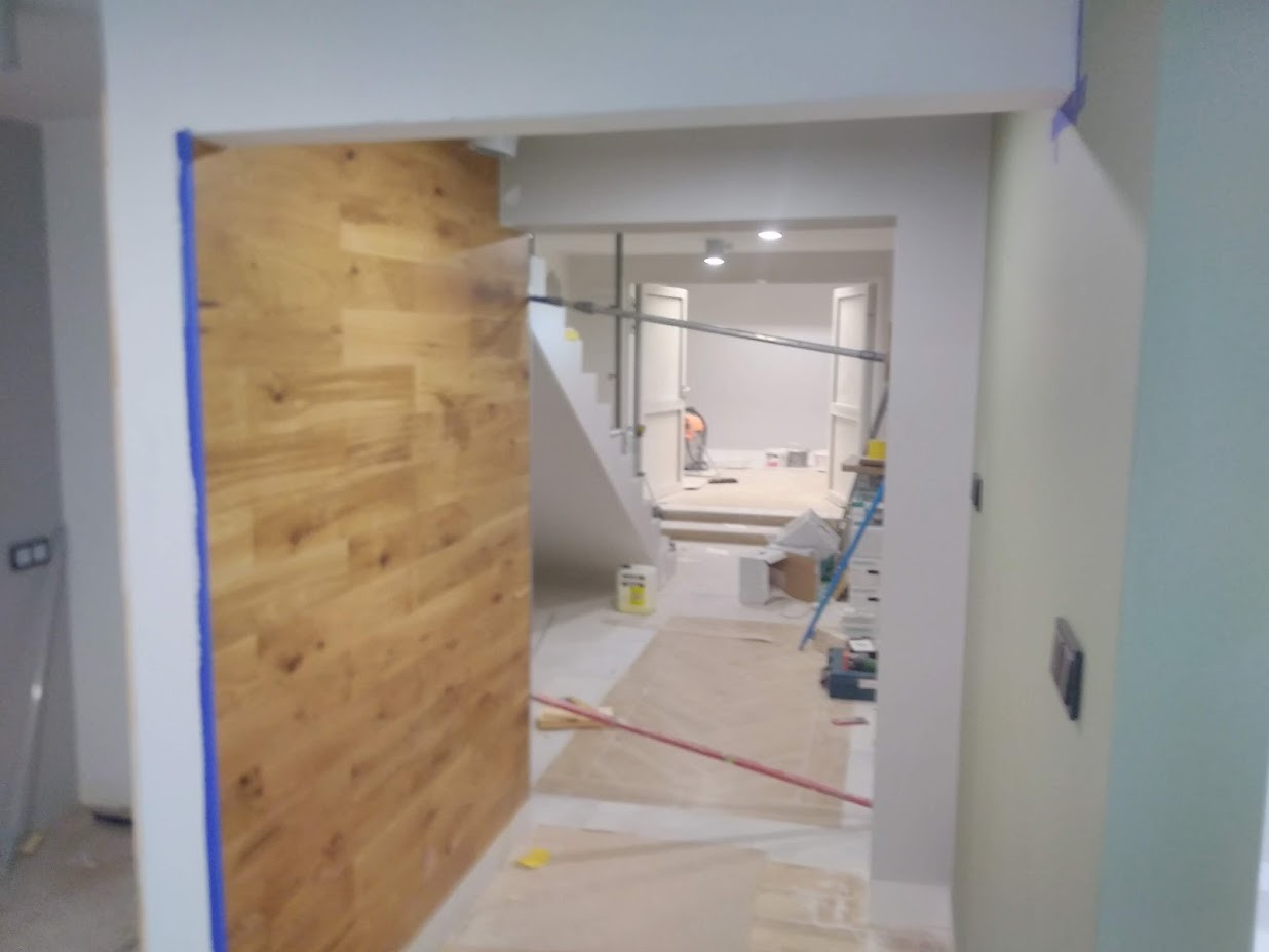 Wnętrze w trakcie remontu: ściana wyłożona drewnianą okładziną, zabezpieczona niebieską taśmą malarską, widoczne narzędzia i materiały budowlane, w tle schody i rusztowanie.