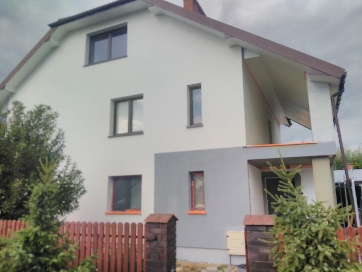 Dwukondygnacyjny dom z częściowo ocieploną elewacją, widoczne różnice w kolorze tynku, fragmenty niezabezpieczonych okien, czerwone ogrodzenie z ceglanymi słupkami.