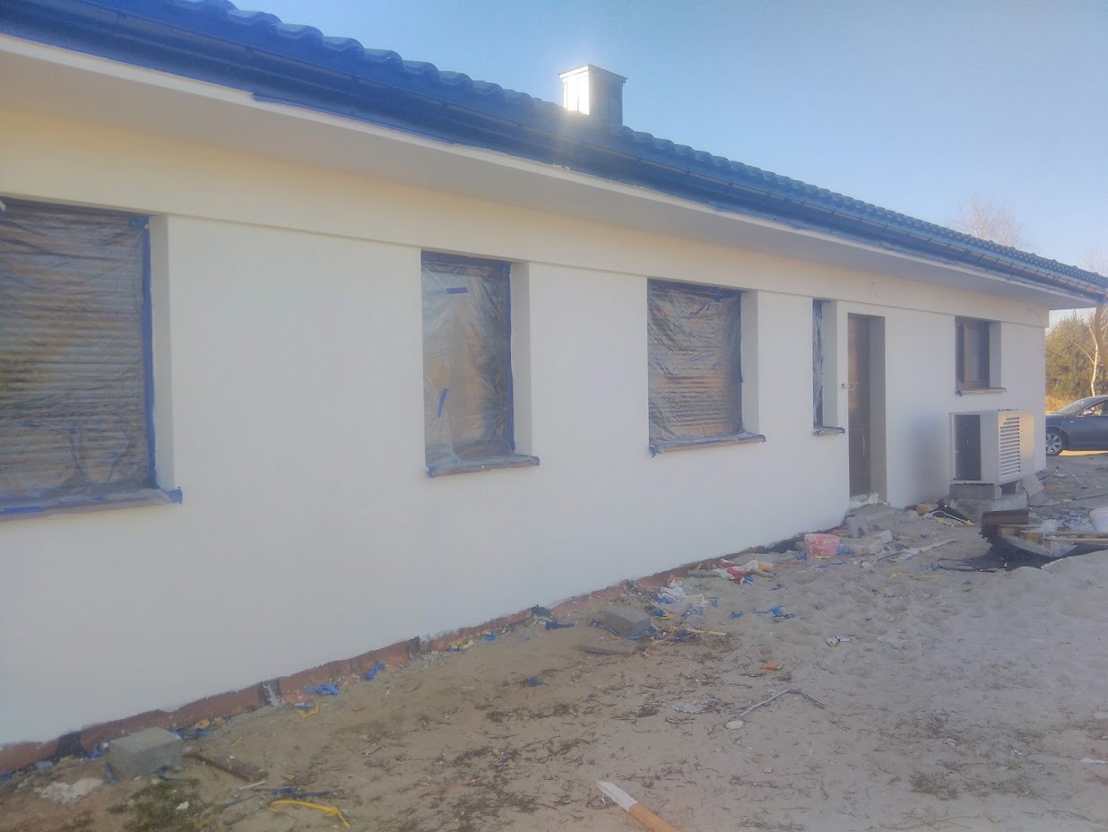 Biała elewacja domu jednorodzinnego w Świdrach po świeżym malowaniu, okna zabezpieczone folią, widoczne ślady prac wykończeniowych na ziemi.