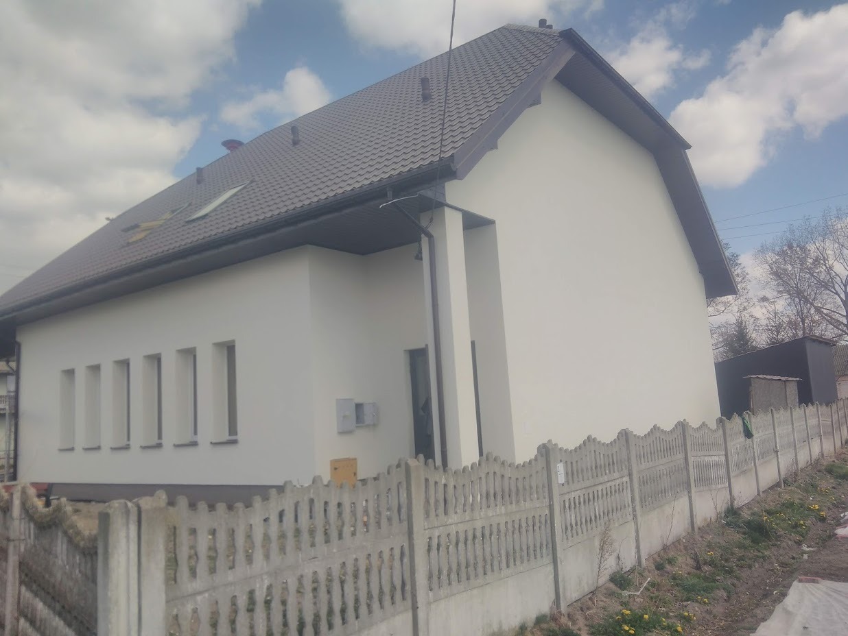 Świeżo pomalowany dom jednorodzinny z szarą dachówką, widok zza betonowego ogrodzenia, biała elewacja, rynna w kolorze dachu.