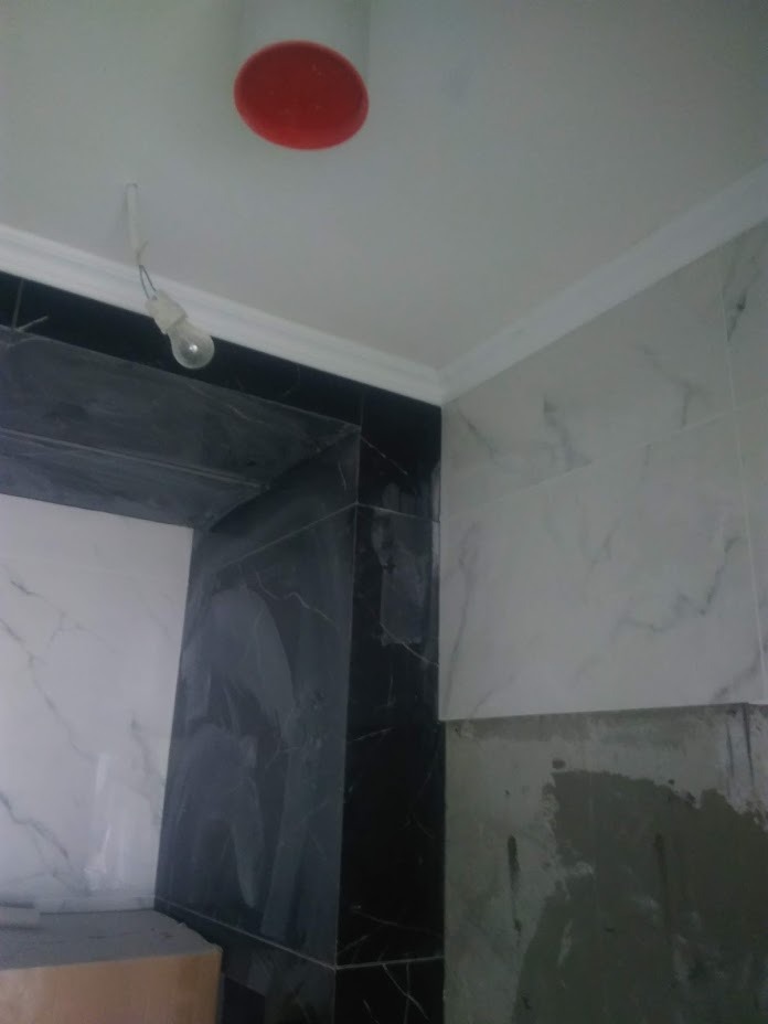 Fragment łazienki w trakcie remontu: czarne i białe marmurowe płytki częściowo ułożone, widoczny surowy tynk na ścianie, biały gzyms pod sufitem i prowizoryczne oświetlenie w postaci żarówki...