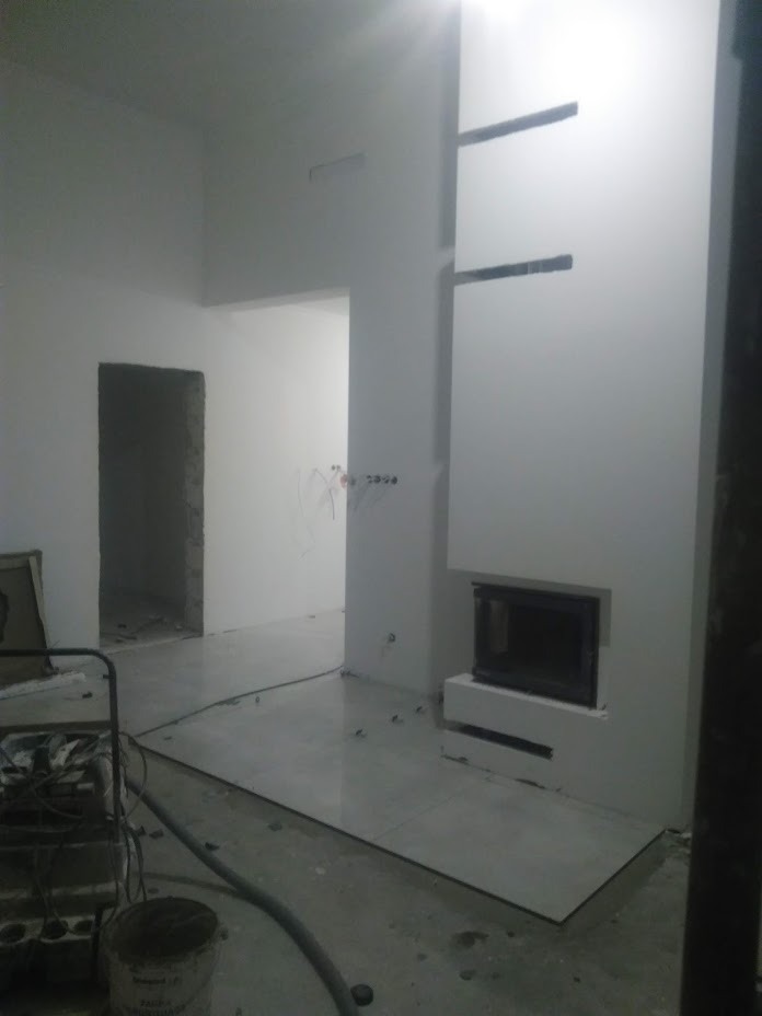 Wnętrze w trakcie remontu: białe ściany, wbudowany kominek z czarnym wkładem, jasna podłoga w płytkach, widoczne kable i narzędzia, surowy otwór drzwiowy w tle.