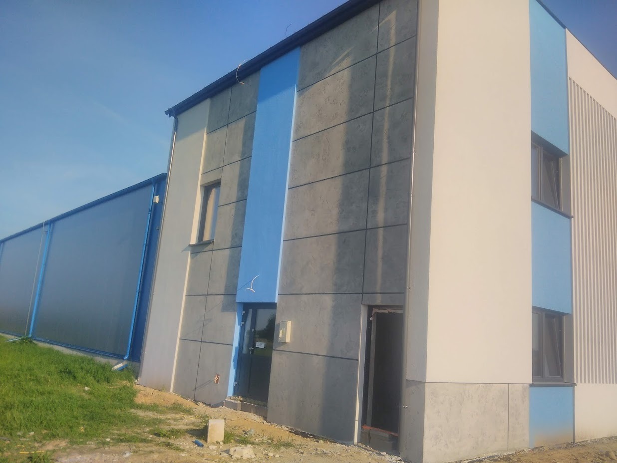 Nowoczesny budynek z elewacją w odcieniach szarości, bieli i błękitu, z widocznymi panelami elewacyjnymi i otwartymi drzwiami wejściowymi.