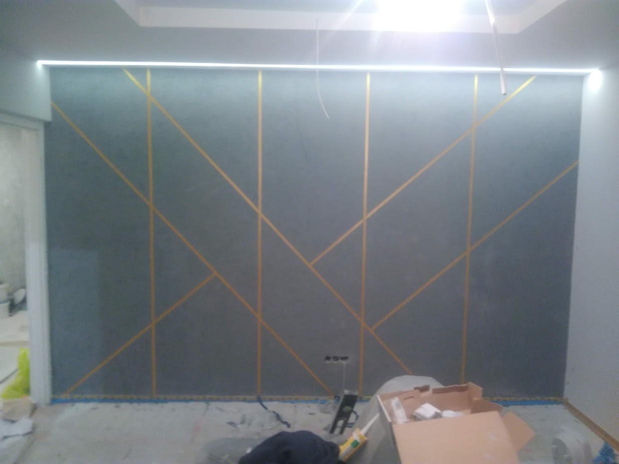 Szara ściana z geometrycznym wzorem z cienkich złotych linii, oświetlenie LED podsufitowe, widoczne ślady prac wykończeniowych na podłodze, kartonowe pudła i narzędzia.