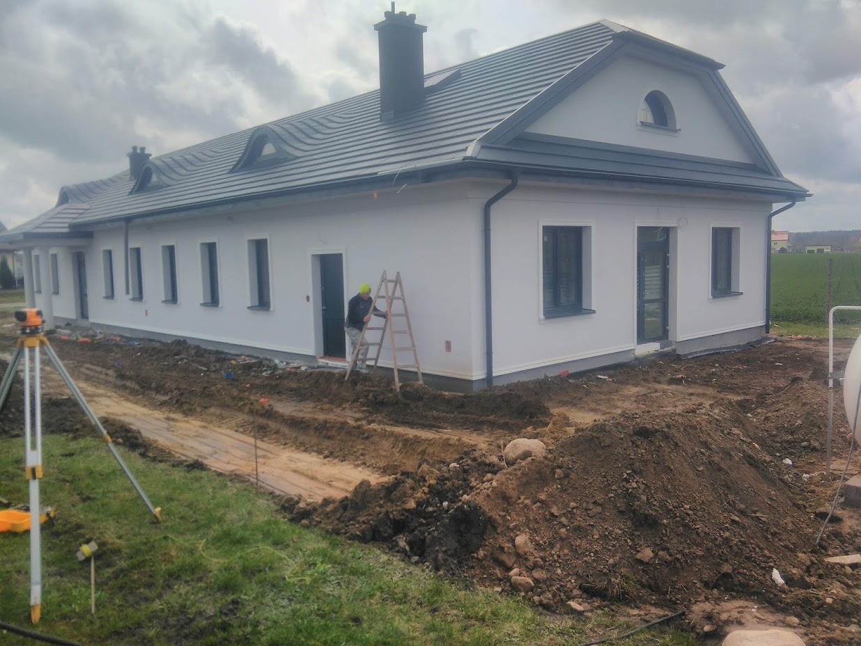 Budynek jednokondygnacyjny z szarą dachówką, białą elewacją i ciemnymi oknami, w trakcie prac wykończeniowych wokół budynku, widoczny pracownik na drabinie i niwelator laserowy.