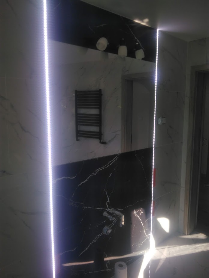 Nowoczesna łazienka z czarno-białymi marmurowymi płytkami, podświetleniem LED wzdłuż krawędzi lustra, grzejnikiem drabinkowym i widocznymi przyłączami hydraulicznymi.