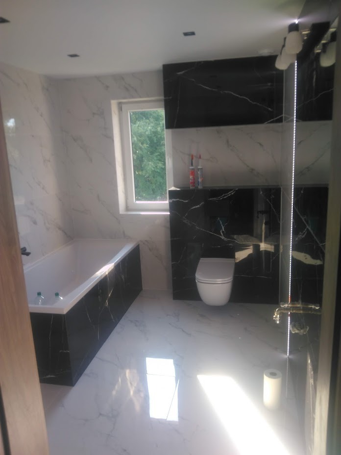 Nowoczesna łazienka z białymi marmurkowymi płytkami podłogowymi i ściennymi, czarną zabudową wanny i szafki pod umywalką, wiszącą toaletą oraz oknem z widokiem na zieleń.