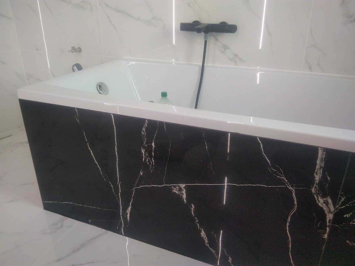 Biała wanna z czarnym panelem imitującym marmur, chromowana bateria, białe marmurowe płytki na ścianach i podłodze, minimalizm.