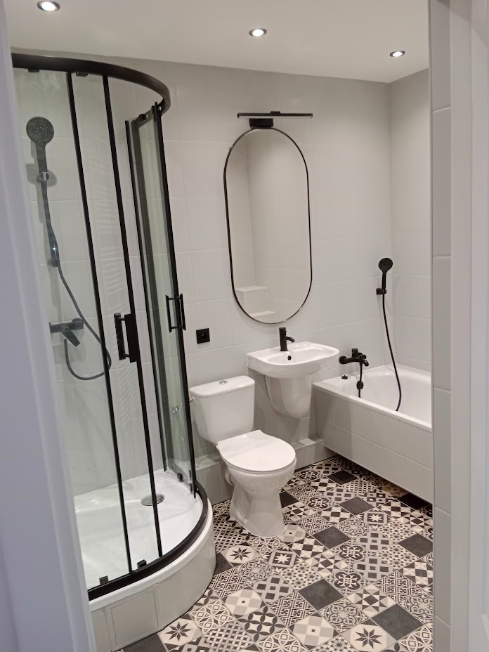 Nowoczesna łazienka z czarnymi akcentami: kabina prysznicowa narożna z czarnymi profilami, owalne lustro z oświetleniem nad umywalką, toaleta i wanna, na podłodze wzorzyste płytki.