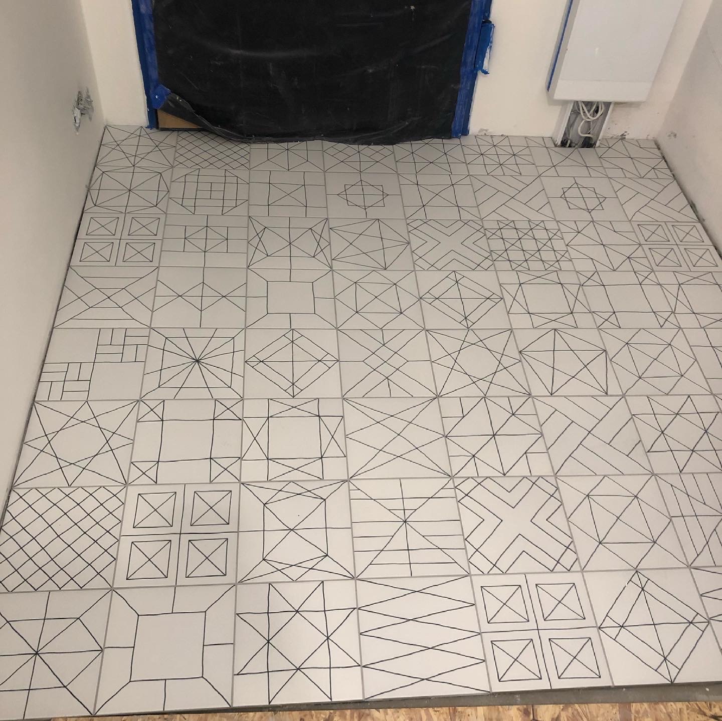 Ułożona podłoga z białych płytek z czarnymi geometrycznymi wzorami w łazience w trakcie remontu; drzwi zabezpieczone folią malarską.