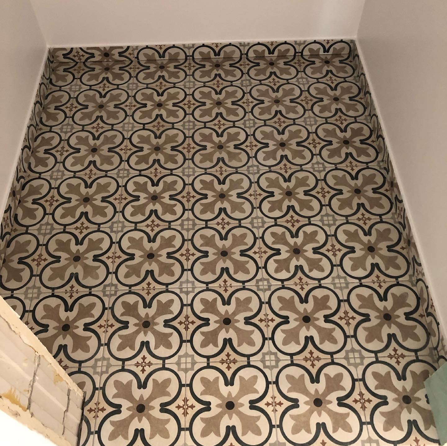 Ułożona podłoga z dekoracyjnych, wzorzystych płytek ceramicznych w odcieniach beżu, brązu i szarości, z widocznym fragmentem niedokończonej ściany po lewej stronie.