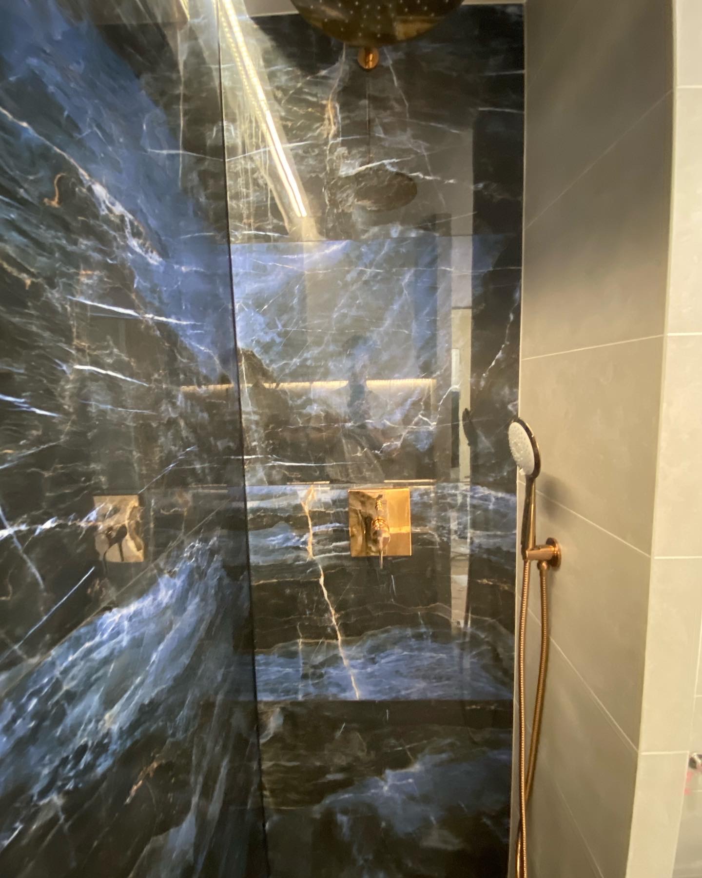 Kabina prysznicowa wyłożona ciemnym marmurem z białymi żyłami, złota armatura, w tym deszczownica i słuchawka prysznicowa.